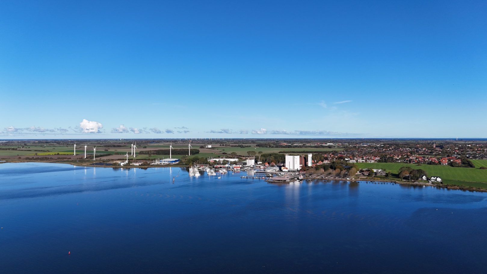 Landschaft mit Wasser, grünen Wiesen und einem kleinen Dorf in der Ferne.